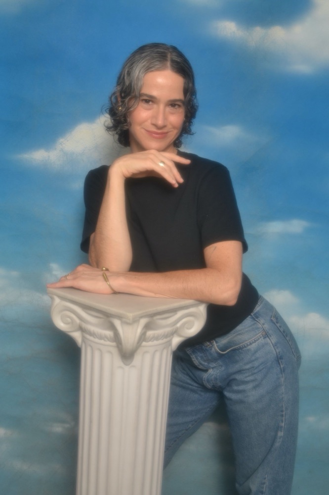 Jaye Fishel leaning on a white classical column against a painted sky backdrop